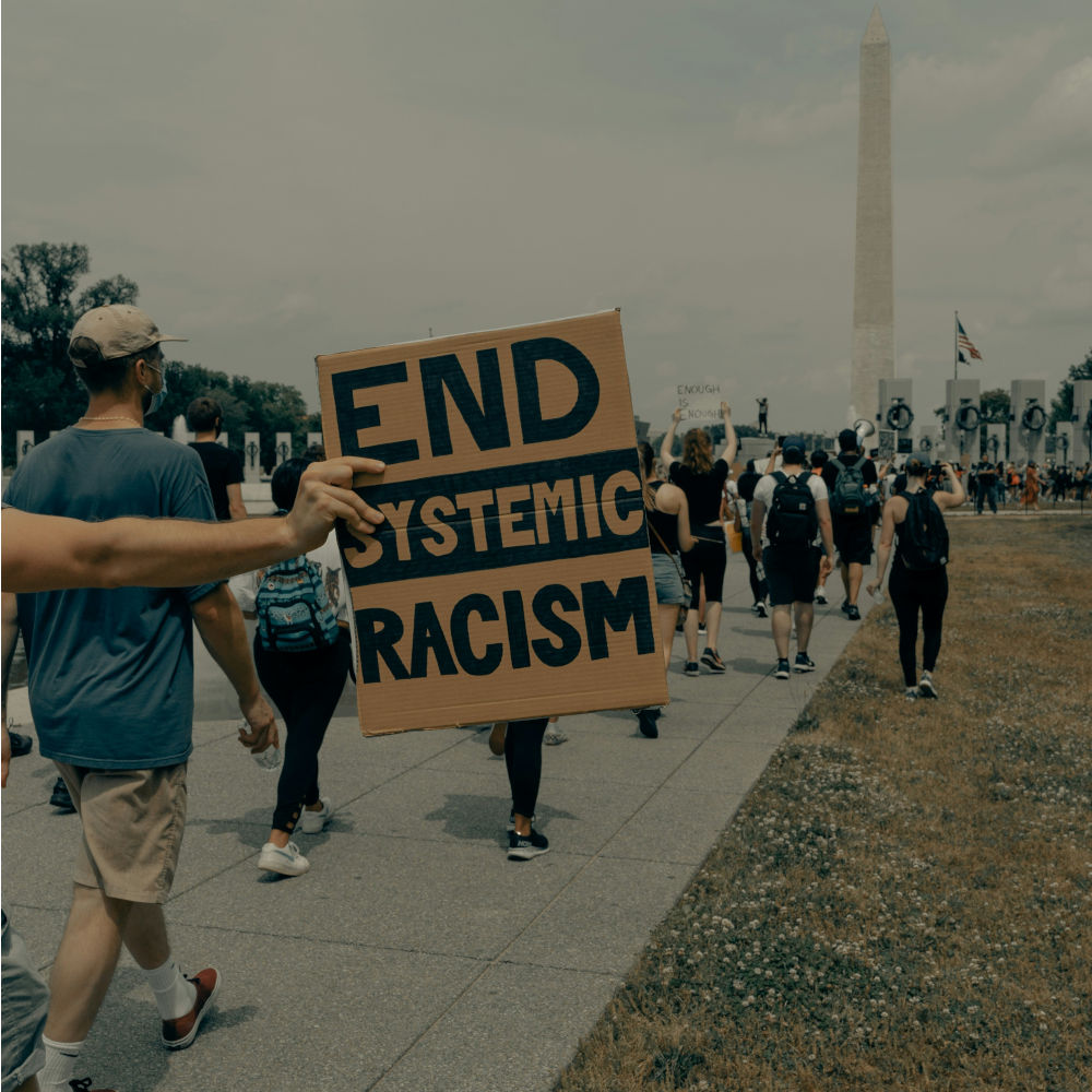 Ein Schild mit der Aufschrift "End systemic racism". Im Hintergrund laufende Menschen.