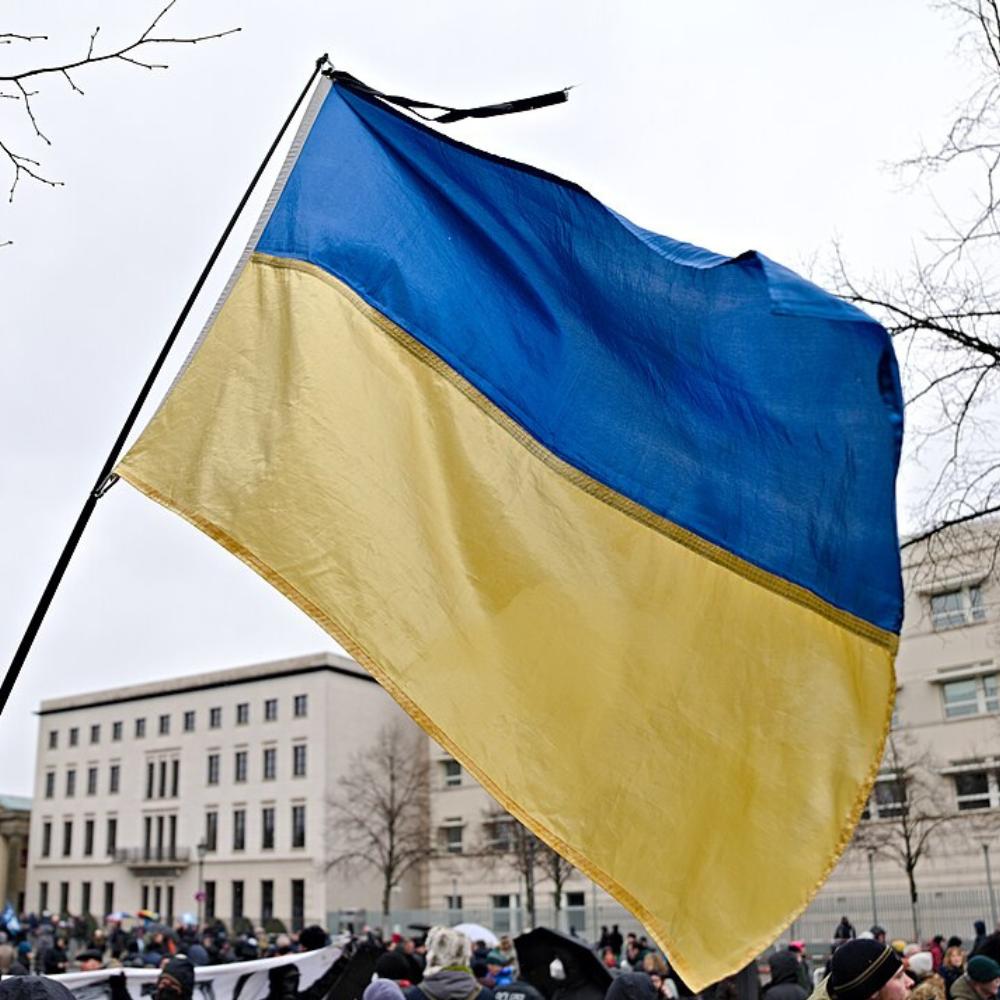 Im Vordergrund ist eine große ukrainische Flagge zu sehen. Im Hintergrund sind viele Menschen, die demonstrieren, sowie ein beiges Gebäude abgebildet.